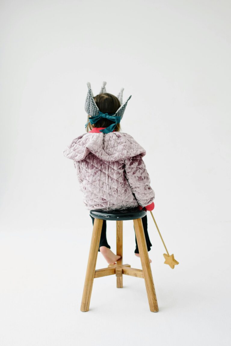 Back view of a child wearing a crown, sitting on a stool with a wand in a white studio.